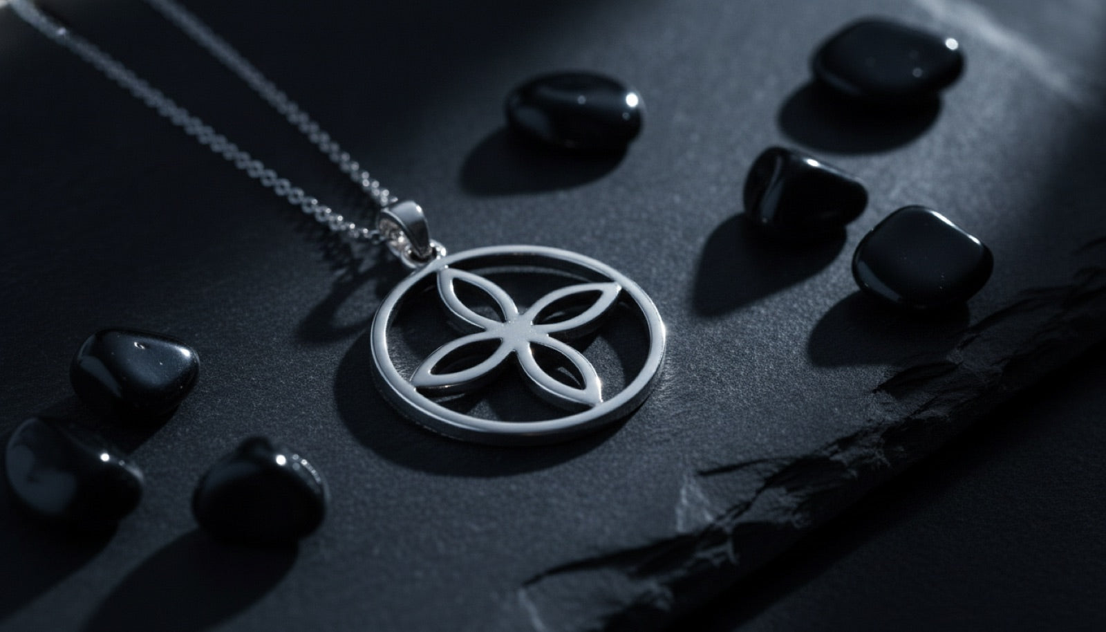 Pendentif argenté au motif nœud de sorcière posé sur de l'ardoise, entouré de pierres d'obsidienne